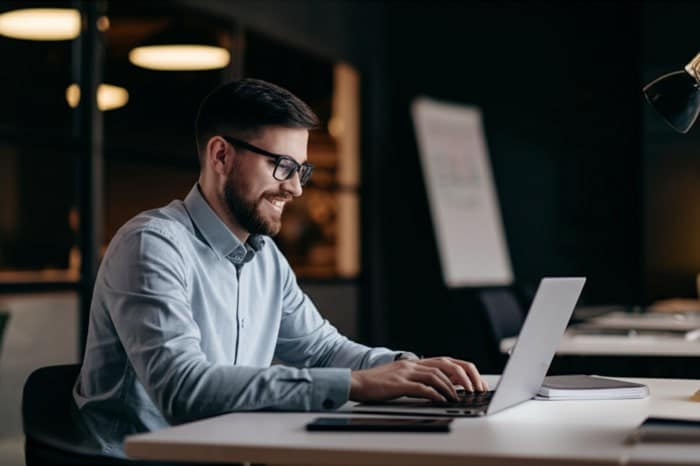 Un lavoratore con gli occhiali e la camicia chiara al lap-top lavora in smart-working serenamente | Qipo